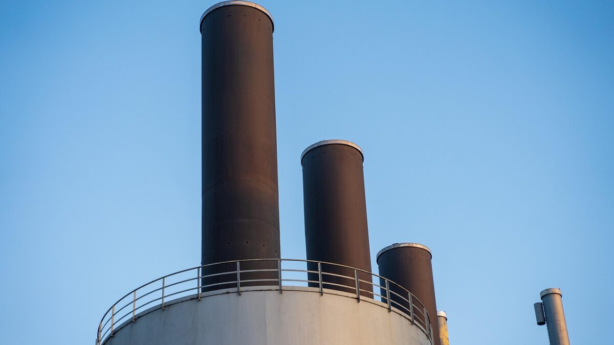 Trois cheminées industrielles sombres sur un grand réservoir blanc, sous un ciel bleu dégagé, avec un petit bâtiment métallique à droite.
