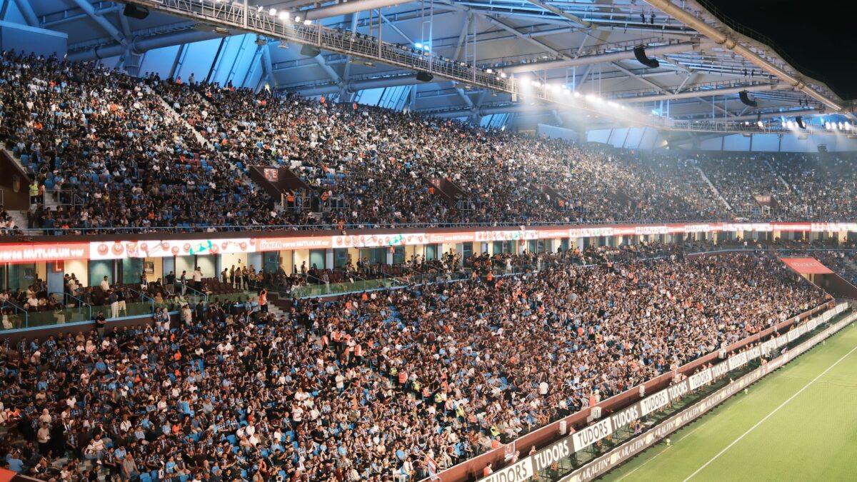 Stade de football bondé la nuit, spectateurs dans les gradins sous éclairage, pelouse verte en bas à droite.