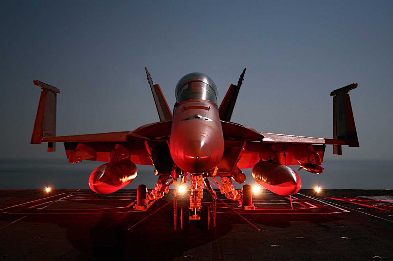 Front view of a fighter jet parked on a dark flight deck at night, lit by red deck lights, with the sea at the horizon behind it.