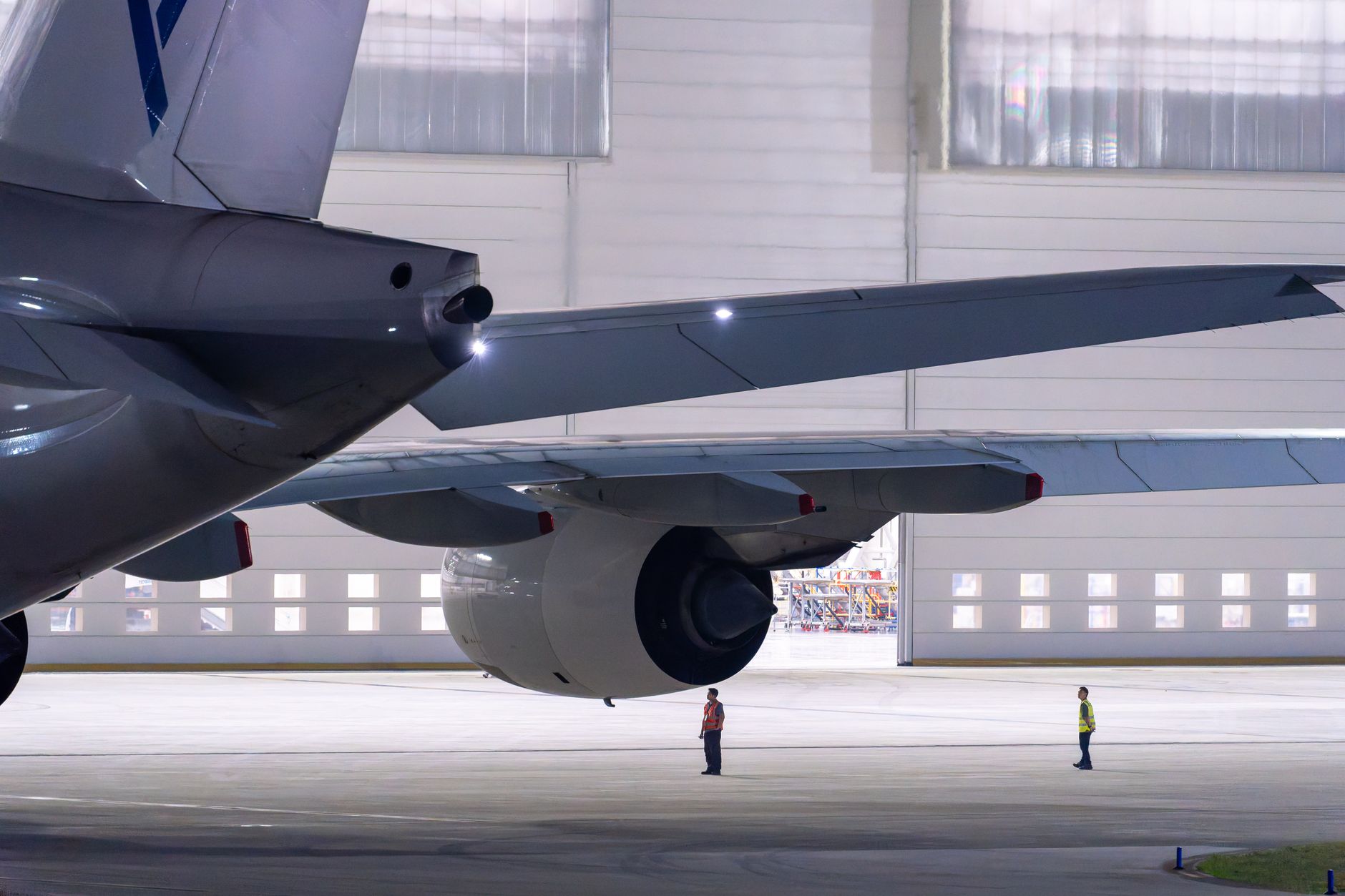 Ground crew personnel standing on the tarmac near a large aircraft wing and jet engine by an open hangar.