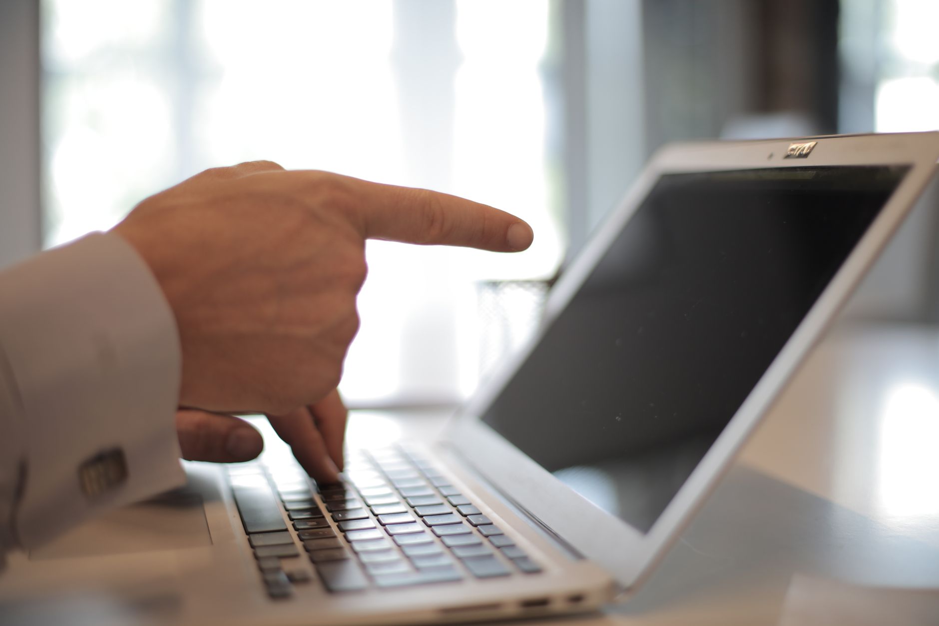 A hand in a shirt cuff points a finger toward the dark screen of a silver laptop on a desk.