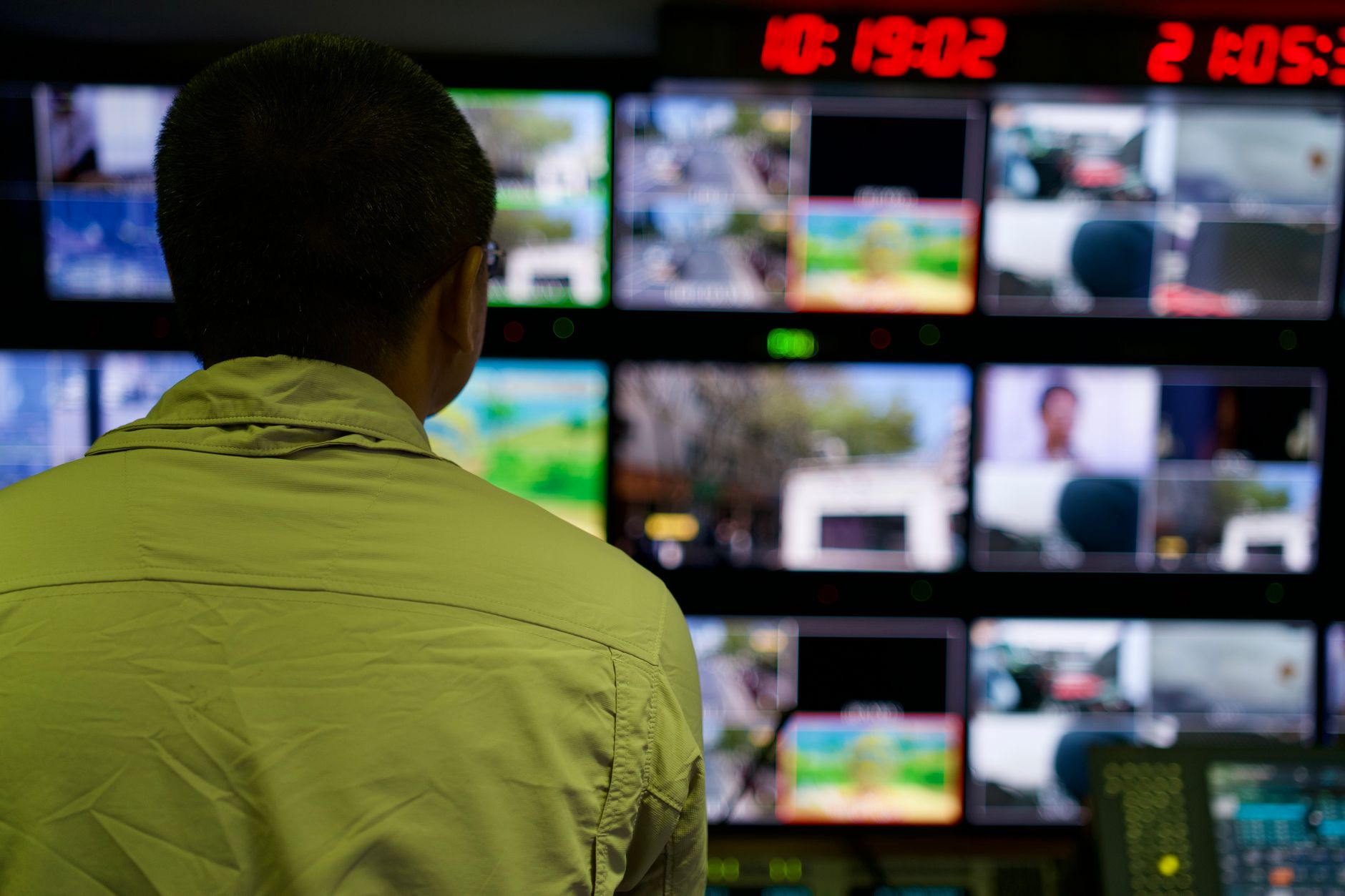 A person viewed from the back observing a wall of television broadcast monitors in a control room.