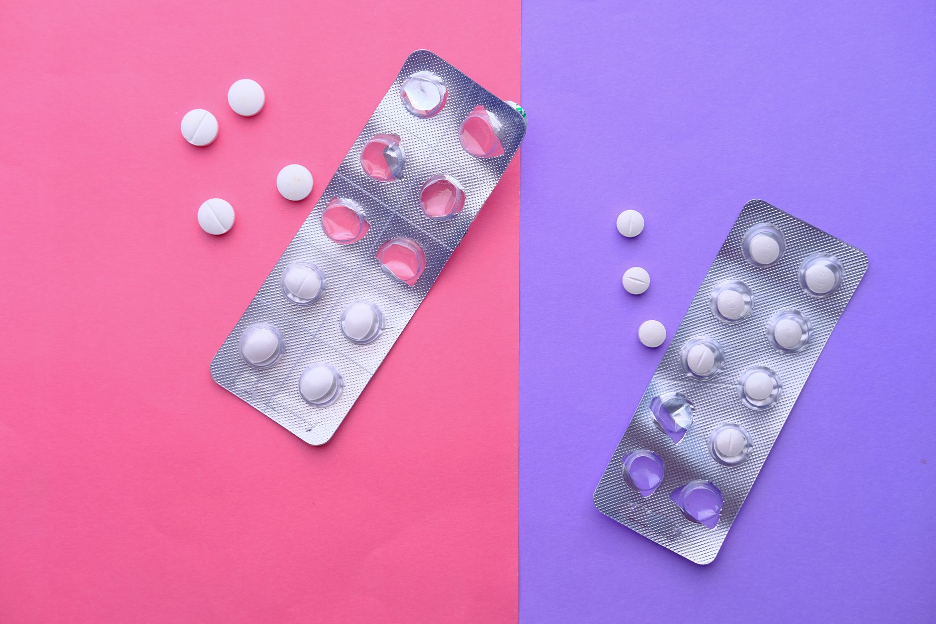 Two silver blister packs of white tablets, partly emptied, with loose pills on a split pink and purple background.