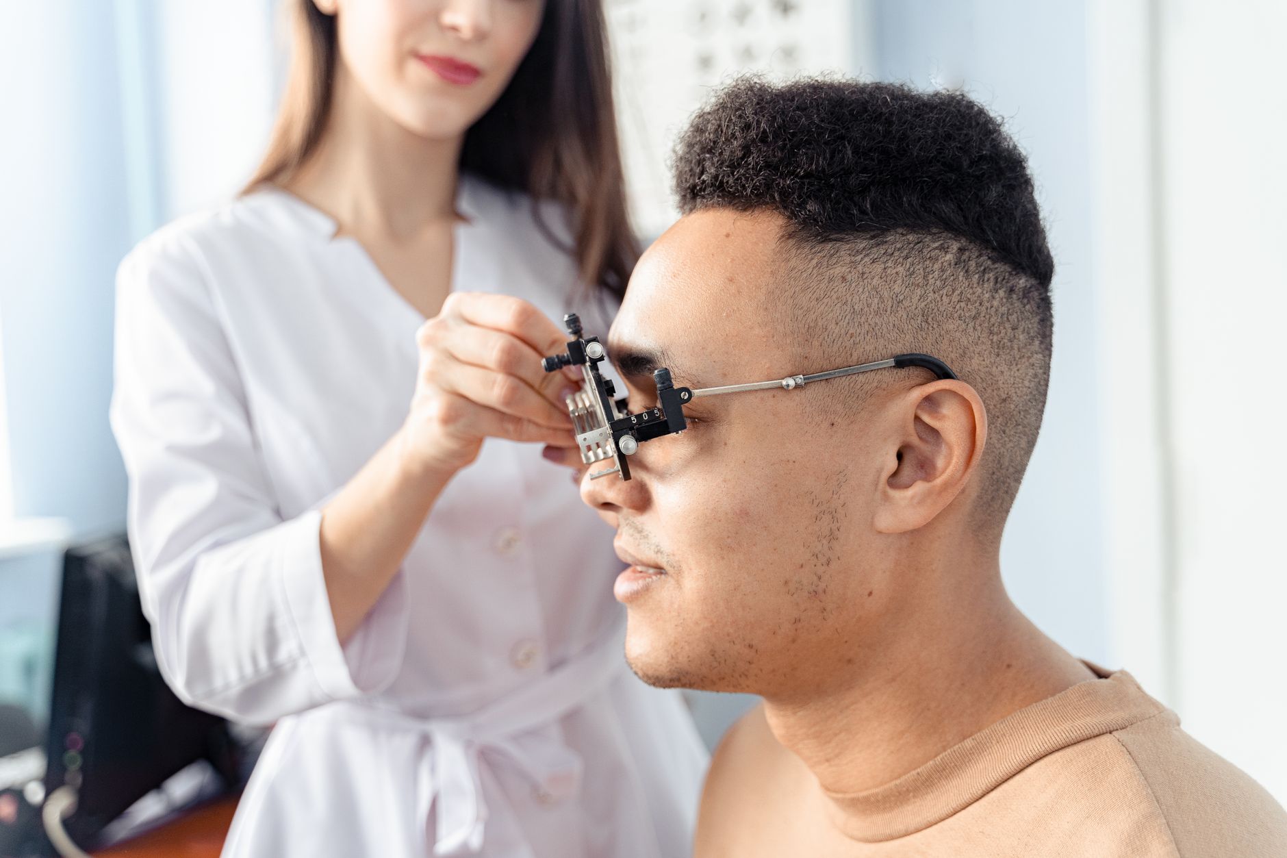 A woman in a white coat adjusts a trial frame on a seated man’s face during an eye exam in a bright clinic.
